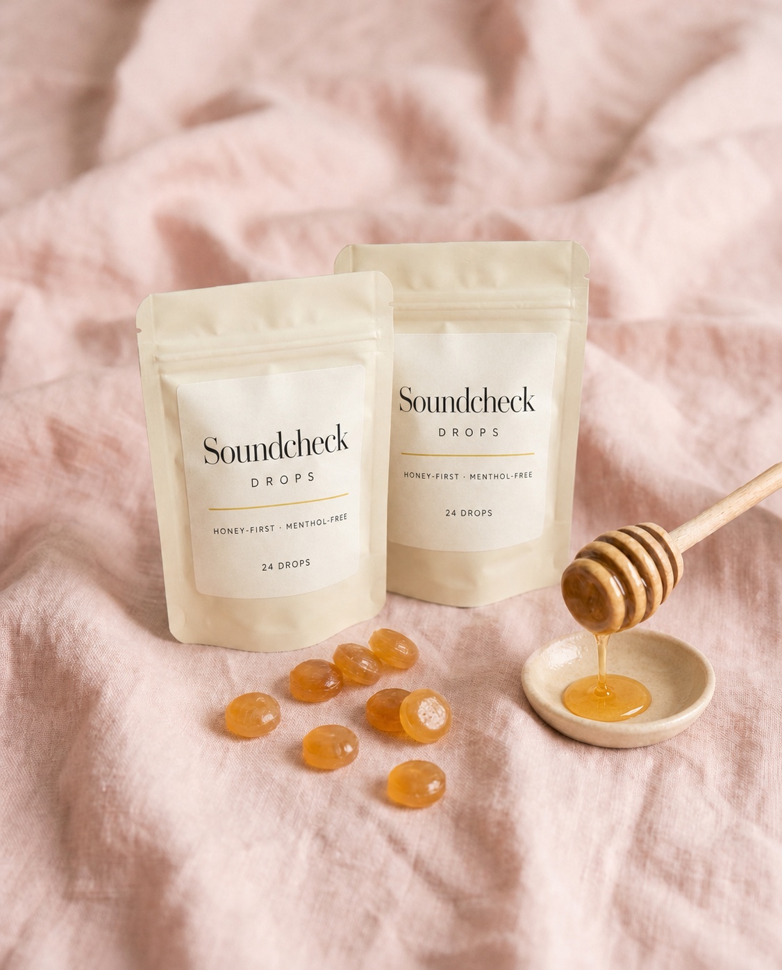 Two labeled pouches of Soundcheck Drops with amber honey drops and a ceramic honey dish on blush linen